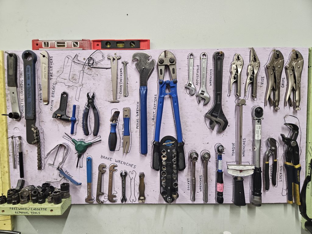 This is a shared tool board. it is painted light purple, like a lavender. The are outlines of tools in black sharpie. There are cassette and freewheel tools. Chain checkers and breakers. Torque wrenches and bolt cutters. A wonderful collection of strong accurate tools.